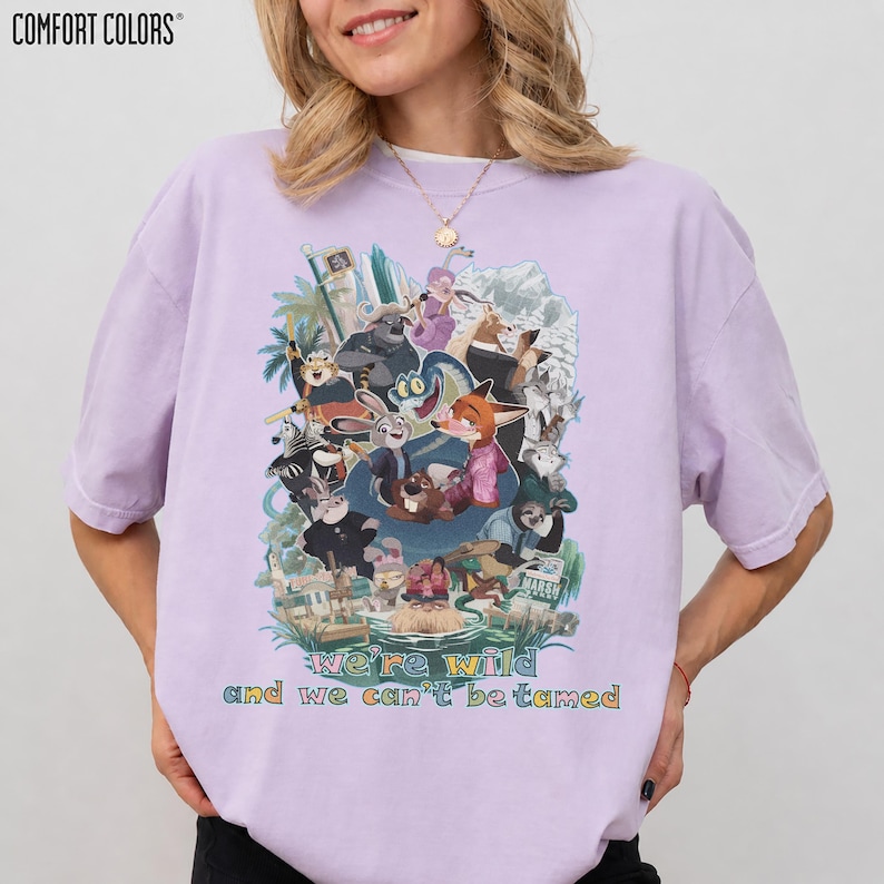May include: A light purple Comfort Colors t-shirt featuring a colorful graphic of various cartoon animals. The text "we're wild and we can't be tamed" is printed below the graphic. The shirt has short sleeves and a crew neck.