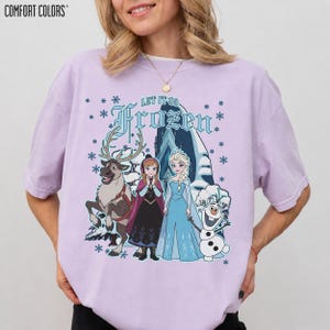 May include: A light purple Comfort Colors t-shirt featuring a graphic from the movie Frozen. The graphic includes the text "Let It Go Frozen" with images of Elsa, Anna, Olaf, and Sven. Snowflakes are scattered around the design.