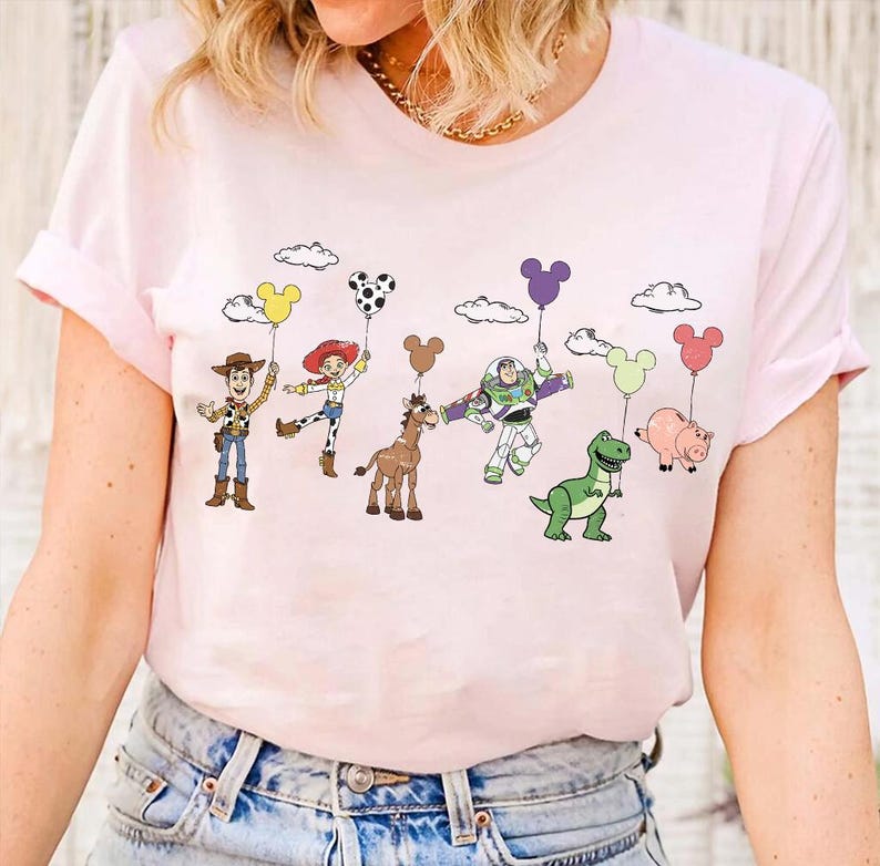 Op de afbeelding: Een lichtroze t-shirt met een kleurrijke afbeelding van stripfiguren die ballonnen vasthouden. De figuren zijn onder meer een cowboy, een cowgirl, een paard, een astronaut, een dinosaurus en een varken. De ballonnen zijn in verschillende kleuren en vormen.