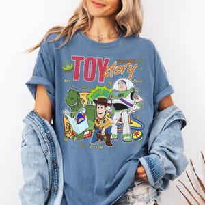 May include: Blue t-shirt featuring the Toy Story logo and characters Rex, Woody, and Buzz Lightyear. The design includes the text "Since 2001" and other graphic elements. The shirt is a comfortable, casual style.
