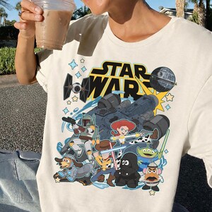 May include: Cream-colored t-shirt featuring a graphic of Star Wars characters and the text "STAR WAR". The design includes a spaceship, the Death Star, and characters like Boba Fett, Woody, and Jessie.