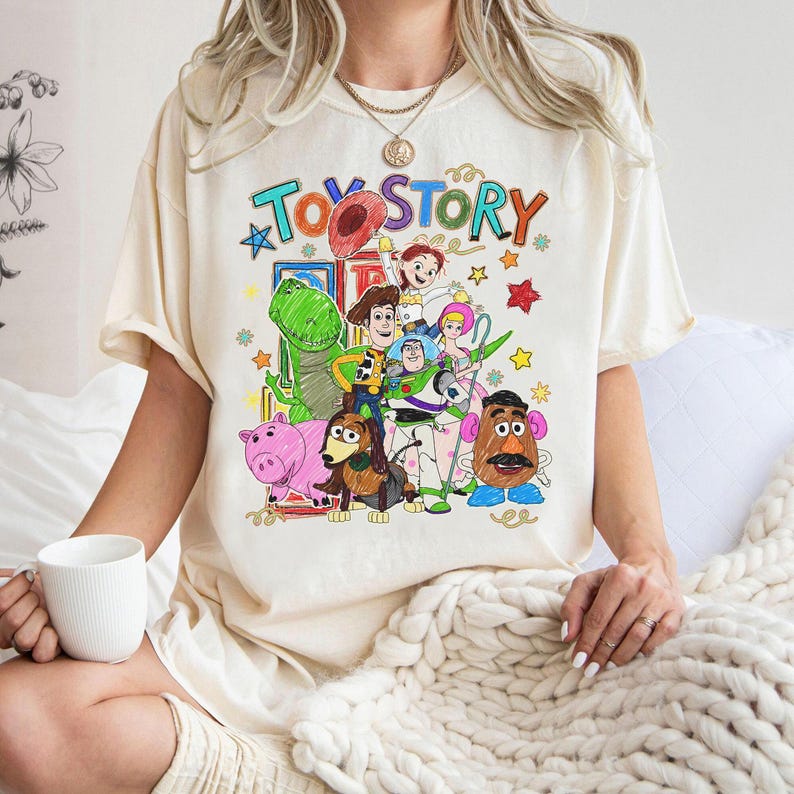 Może przedstawiać: Kremowa koszulka z kolorową grafiką postaci z filmu animowanego "Toy Story". Projekt zawiera napis "Toy Story" w zabawnej czcionce, a także obrazy Woody'ego, Buzza Astrala i innych postaci.