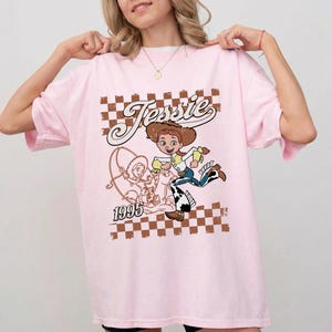 May include: Pink t-shirt featuring a cartoon graphic of Jessie from Toy Story. The design includes the name "Jessie" in a vintage font, a checkered background, and the year "1995". The graphic is in shades of brown, yellow, blue, and white.