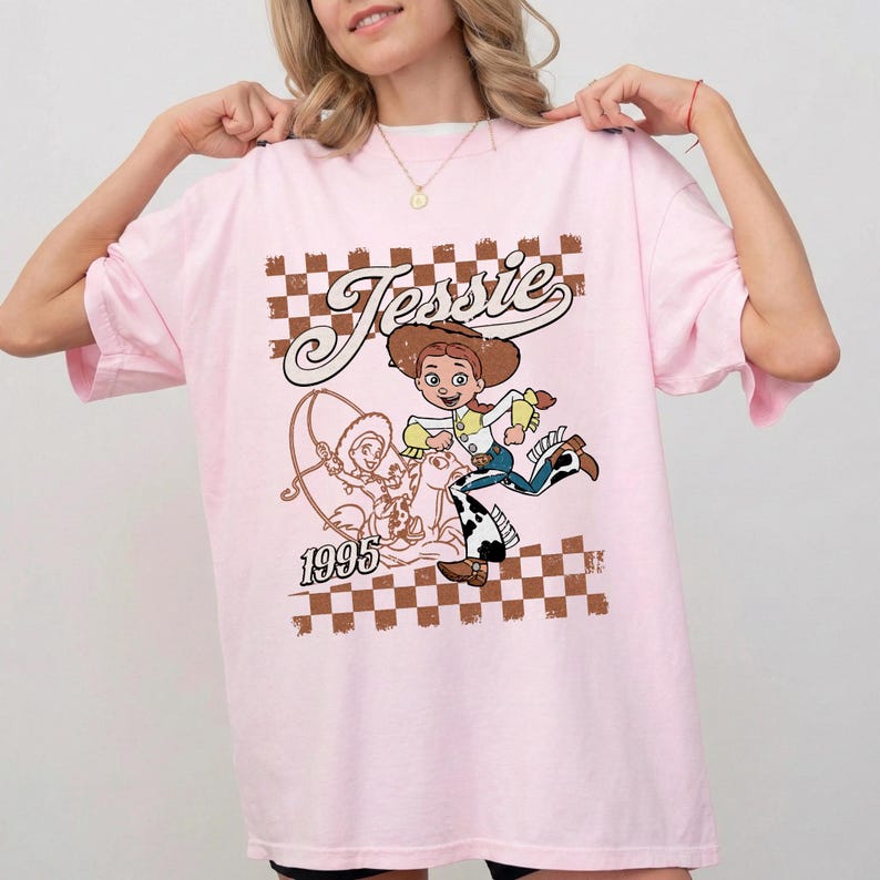 May include: Pink t-shirt featuring a cartoon graphic of Jessie from Toy Story. The design includes the name "Jessie" in a vintage font, a checkered background, and the year "1995". The graphic is in shades of brown, yellow, blue, and white.