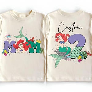 May include: Cream-colored toddler t-shirts with a mermaid theme. The front features the word "MOM" with a mermaid illustration. The back has a mermaid illustration with the number "2" and the word "Custom".