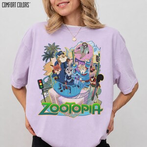 Puede incluir: Una camiseta Comfort Colors de color lila claro con un gr&aacute;fico colorido de la pel&iacute;cula Zootopia. El dise&ntilde;o presenta personajes de animales y la palabra "Zootopia" en verde y dorado.