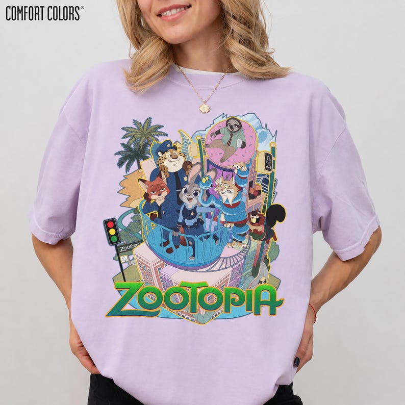 Puede incluir: Una camiseta Comfort Colors de color lila claro con un gr&aacute;fico colorido de la pel&iacute;cula Zootopia. El dise&ntilde;o presenta personajes de animales y la palabra "Zootopia" en verde y dorado.