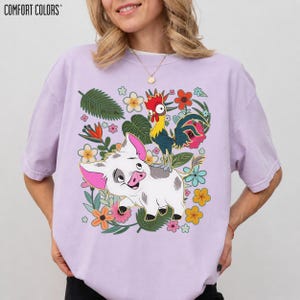 Puede incluir: Camiseta Comfort Colors de color lila claro con un gr&aacute;fico colorido de un cerdo y un gallo rodeados de flores y hojas. El cerdo es blanco con orejas y hocico rosas. El gallo es amarillo, rojo y azul.