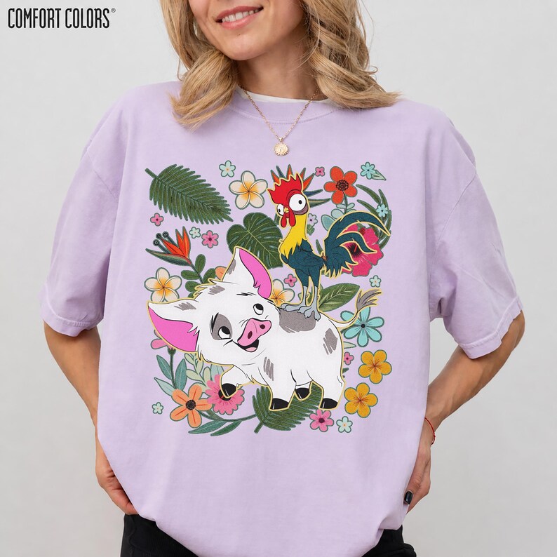 Puede incluir: Camiseta Comfort Colors de color lila claro con un gr&aacute;fico colorido de un cerdo y un gallo rodeados de flores y hojas. El cerdo es blanco con orejas y hocico rosas. El gallo es amarillo, rojo y azul.