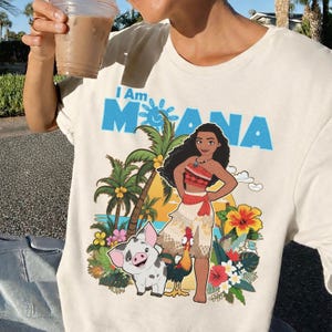May include: Cream-colored t-shirt featuring the text "I Am Moana" in blue. The graphic includes an illustration of Moana, a pig, a rooster, palm trees, and tropical flowers. The shirt is a casual wear item.