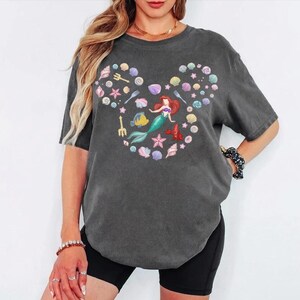 May include: Dark gray t-shirt featuring a colorful graphic of a mermaid and sea creatures arranged in a Mickey Mouse head shape. The design includes seashells, starfish, and a fish. The shirt has short sleeves and a crew neck.