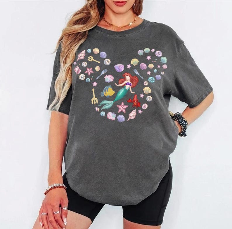 May include: Dark gray t-shirt featuring a colorful graphic of a mermaid and sea creatures arranged in a Mickey Mouse head shape. The design includes seashells, starfish, and a fish. The shirt has short sleeves and a crew neck.