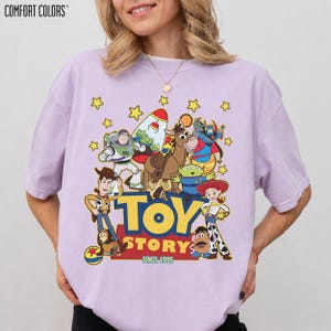 May include: A light purple Comfort Colors t-shirt featuring a graphic of Toy Story characters, including Woody, Buzz Lightyear, and Jessie. The words "TOY STORY" and "SINCE 1995" are printed in yellow and red.