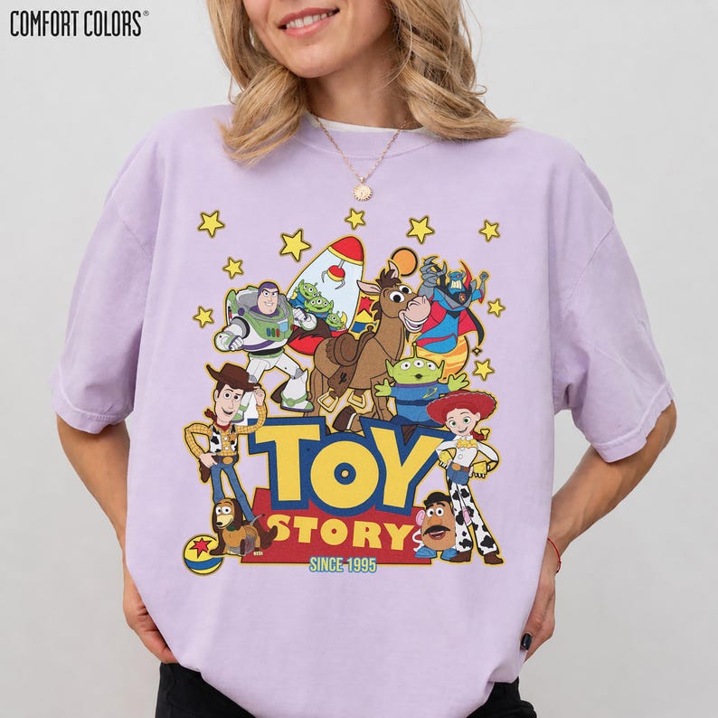 May include: A light purple Comfort Colors t-shirt featuring a graphic of Toy Story characters, including Woody, Buzz Lightyear, and Jessie. The words "TOY STORY" and "SINCE 1995" are printed in yellow and red.