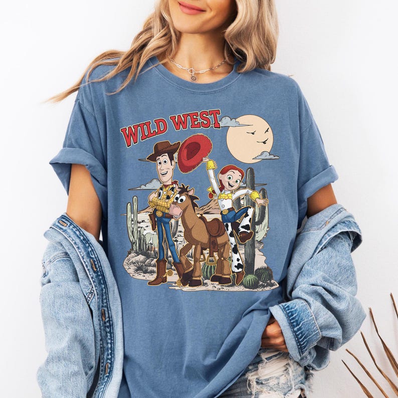 Pu&ograve; includere: T-shirt blu con grafica Wild West con i personaggi dei cartoni animati Woody, Jessie e un cavallo. Il design include un paesaggio desertico con cactus, un sole e le parole "Wild West" in rosso.