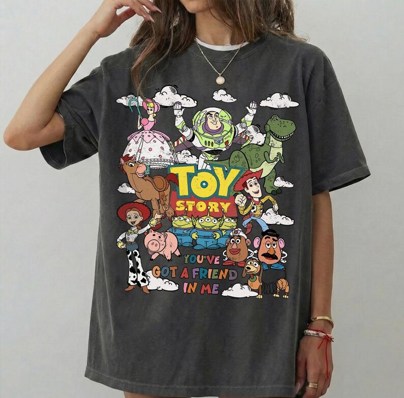 Op de afbeelding: Donkergrijze t-shirt met een kleurrijke afbeelding van personages uit de film "Toy Story". Het ontwerp bevat Woody, Buzz Lightyear en andere personages, met de tekst "Toy Story" en "You've Got a Friend in Me."