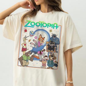 May include: A cream-colored short-sleeve t-shirt featuring a large rectangular graphic print with the word Zootopia in green text. The illustration displays various animated characters including a fox, a rabbit, a sloth, and a snake in a colorful scene.