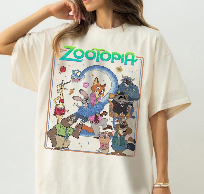 May include: A cream-colored short-sleeve t-shirt featuring a large rectangular graphic print with the word Zootopia in green text. The illustration displays various animated characters including a fox, a rabbit, a sloth, and a snake in a colorful scene.