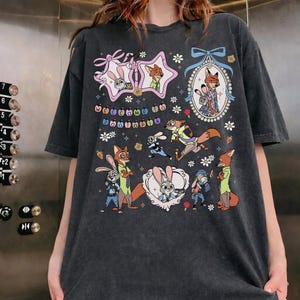 May include: A charcoal gray short-sleeve t-shirt featuring a graphic print of characters from Zootopia. The design includes multiple illustrations of a rabbit and a fox, decorative frames, small flowers, and the text Welcome to Zootopia.