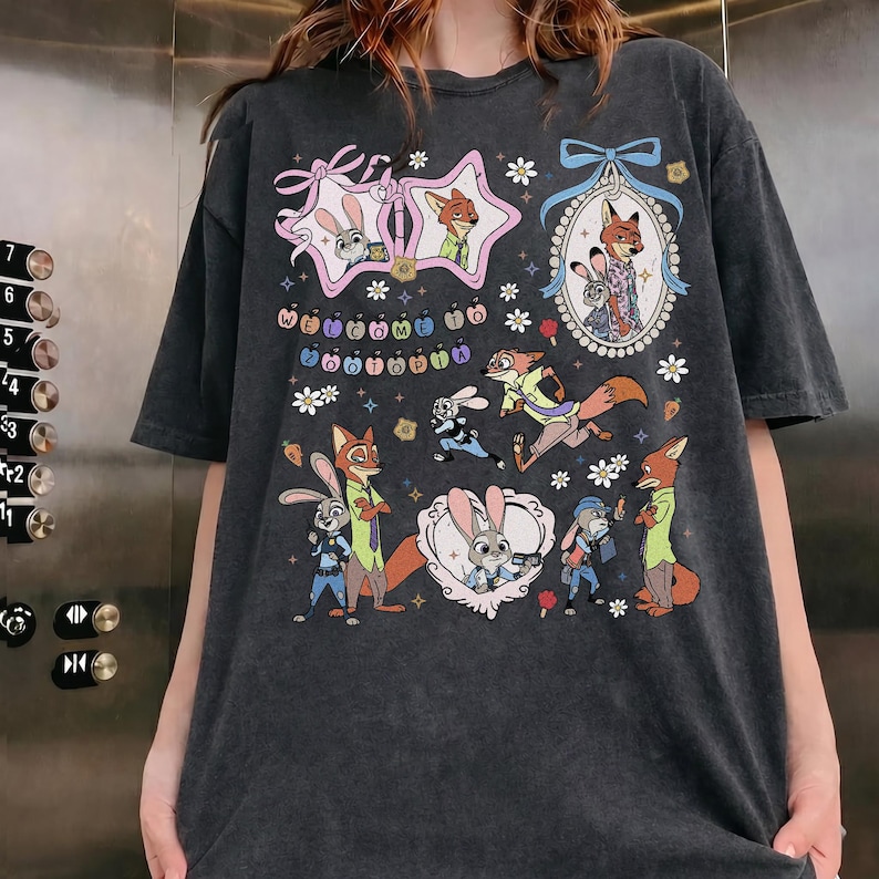 May include: A charcoal gray short-sleeve t-shirt featuring a graphic print of characters from Zootopia. The design includes multiple illustrations of a rabbit and a fox, decorative frames, small flowers, and the text Welcome to Zootopia.
