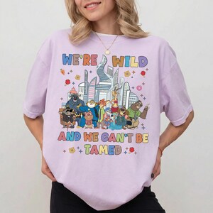 K&ouml;nnte beinhalten: Ein hellviolettes T-Shirt mit einer bunten Grafik verschiedener animierter Tierfiguren aus Zootopia vor einer Stadtsilhouette. Der Text WE'RE WILD AND WE CAN'T BE TAMED ist in mehrfarbigen Buchstaben &uuml;ber und unter den Charakteren aufgedruckt.