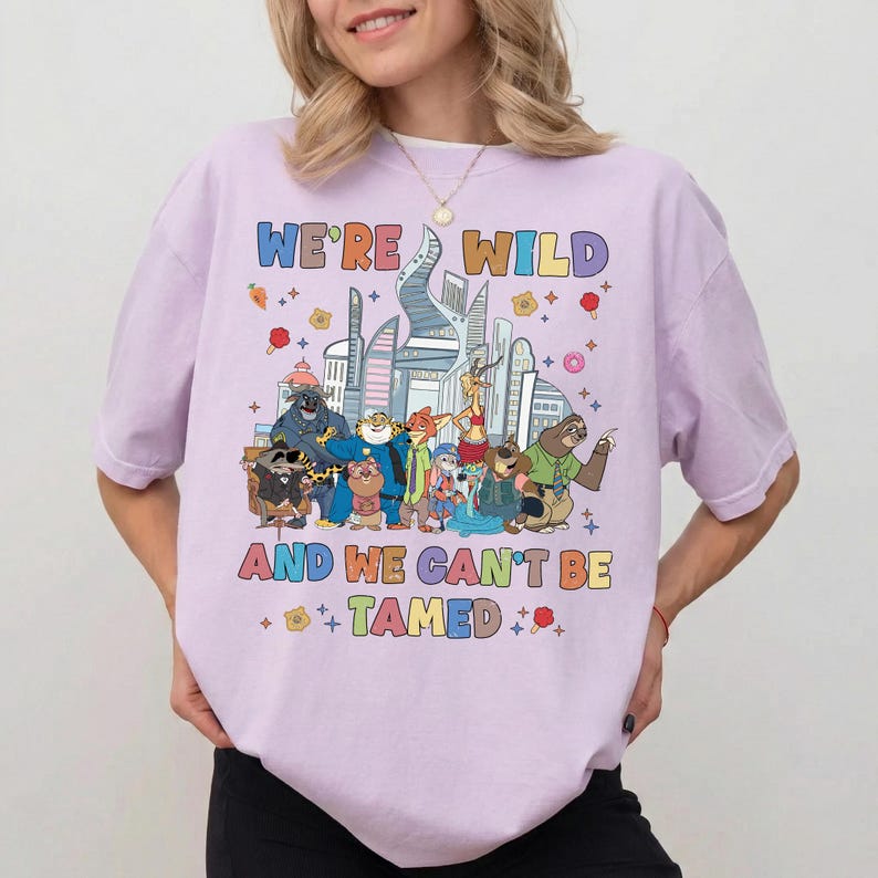 K&ouml;nnte beinhalten: Ein hellviolettes T-Shirt mit einer bunten Grafik verschiedener animierter Tierfiguren aus Zootopia vor einer Stadtsilhouette. Der Text WE'RE WILD AND WE CAN'T BE TAMED ist in mehrfarbigen Buchstaben &uuml;ber und unter den Charakteren aufgedruckt.
