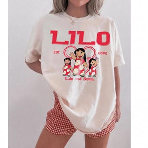 Puede incluir: Una camiseta de manga corta color hueso con un estampado gr&aacute;fico rojo. El texto dice LILO en la parte superior, EST. 2002 en el medio y Lilo and Stitch en la parte inferior. El gr&aacute;fico muestra tres personajes de Lilo con vestidos rojos florales.