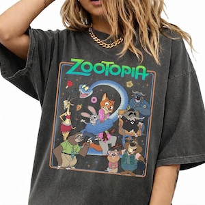 May include: A charcoal gray short-sleeve t-shirt featuring a colorful graphic print of Zootopia characters including Judy Hopps, Nick Wilde, and Flash. The design is enclosed in a rectangular border with the text Zootopia at the top in green.