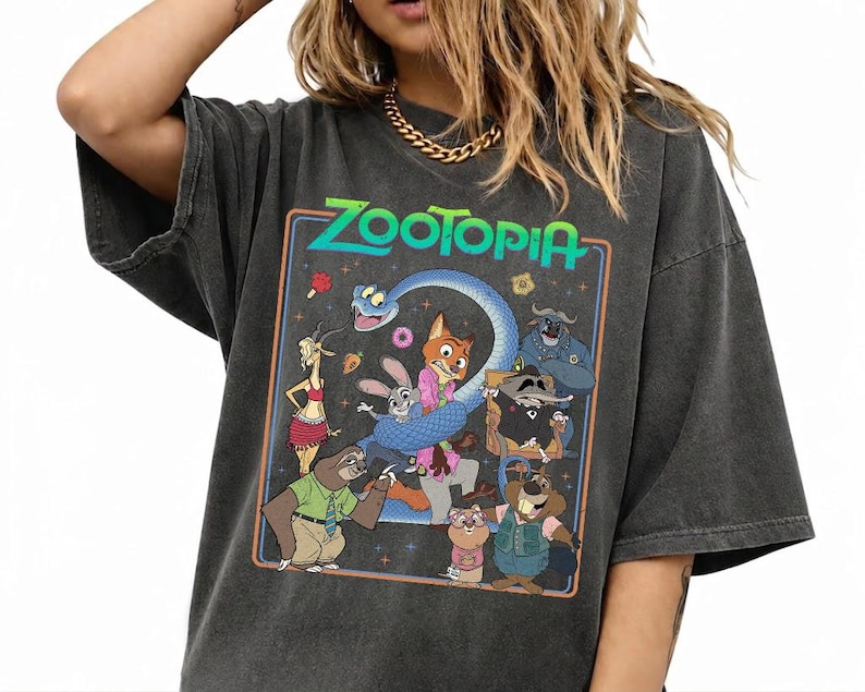 May include: A charcoal gray short-sleeve t-shirt featuring a colorful graphic print of Zootopia characters including Judy Hopps, Nick Wilde, and Flash. The design is enclosed in a rectangular border with the text Zootopia at the top in green.