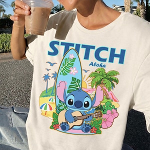 May include: Cream-colored t-shirt featuring a graphic of Stitch playing a guitar in front of a surfboard, palm trees, and flowers. The word "STITCH" is printed above the graphic in blue, with "Aloha" below. The shirt is a casual wear item.
