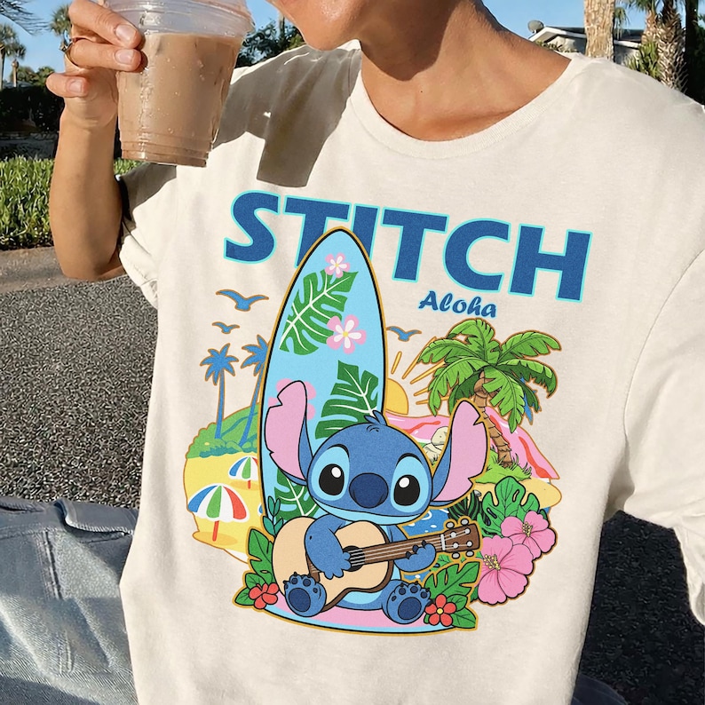 May include: Cream-colored t-shirt featuring a graphic of Stitch playing a guitar in front of a surfboard, palm trees, and flowers. The word "STITCH" is printed above the graphic in blue, with "Aloha" below. The shirt is a casual wear item.