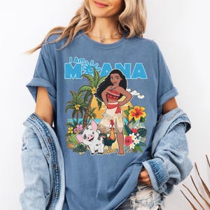 May include: Blue t-shirt featuring the phrase "I Am Moana" above a graphic of Moana, Pua the pig, and Hei Hei the rooster. The design includes palm trees, flowers, and a sunset background. The shirt is a casual style.