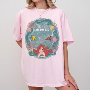 May include: A light pink t-shirt featuring a circular graphic of Ariel from The Little Mermaid. The graphic includes Ariel, Flounder, Sebastian, and the text "The Little Mermaid" in a teal frame with coral and seashells.