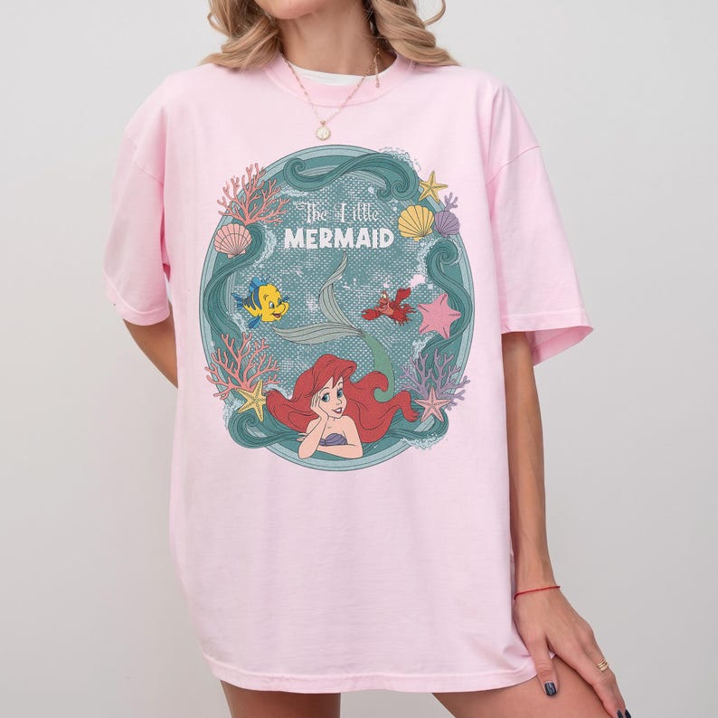 May include: A light pink t-shirt featuring a circular graphic of Ariel from The Little Mermaid. The graphic includes Ariel, Flounder, Sebastian, and the text "The Little Mermaid" in a teal frame with coral and seashells.