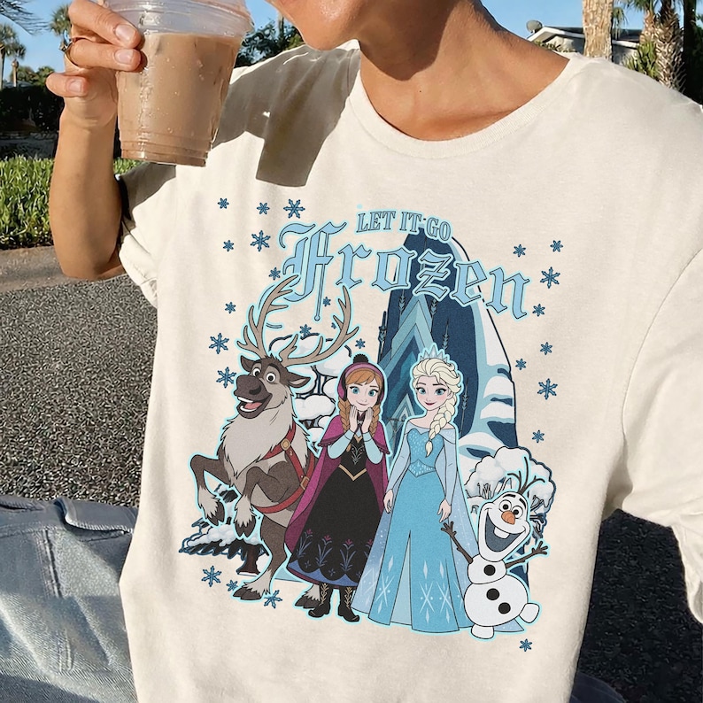 May include: Cream-colored t-shirt featuring a graphic from the movie "Frozen." The design includes the text "LET IT GO Frozen" with images of characters Anna, Elsa, Olaf, and Sven. Blue snowflakes surround the characters.