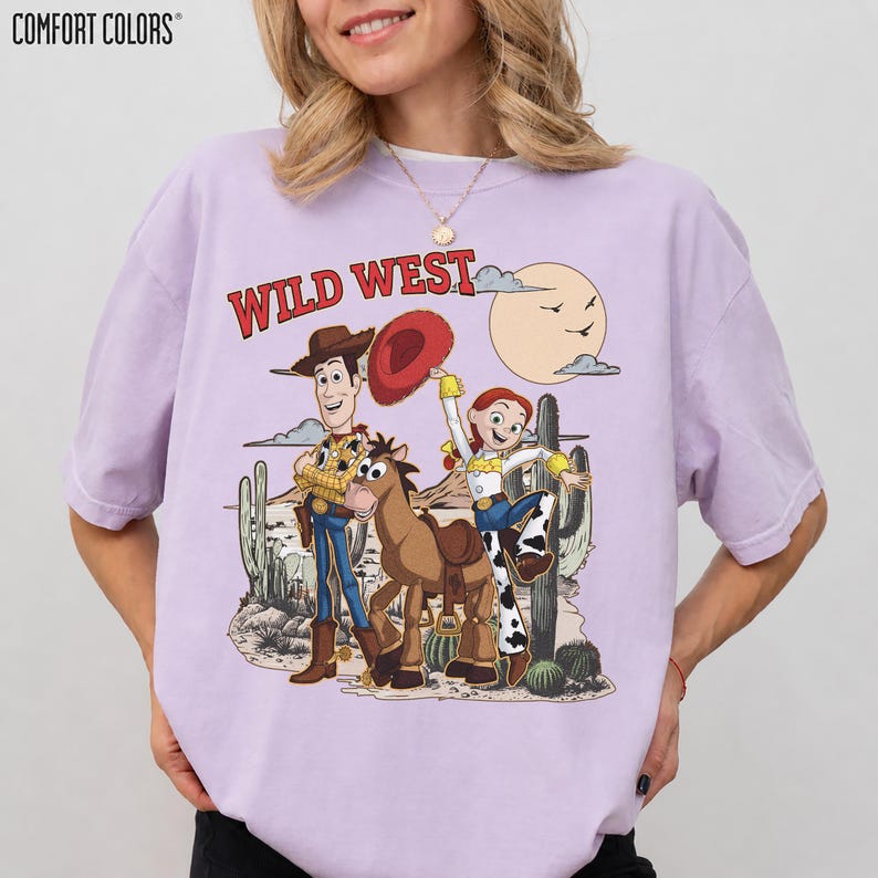 Pu&ograve; includere: T-shirt Comfort Colors lilla chiaro con grafica Wild West. La grafica include personaggi dei cartoni animati, un cavallo, cactus e un sole. Il testo "WILD WEST" &egrave; in rosso sopra i personaggi. La maglietta ha una vestibilit&agrave; ampia.