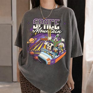 May include: Dark grey t-shirt with a colourful graphic of the Space Mountain ride featuring Mickey Mouse, Donald Duck, Goofy, and Pluto. The shirt has the words "SPACE Mountain" in a purple and white design.