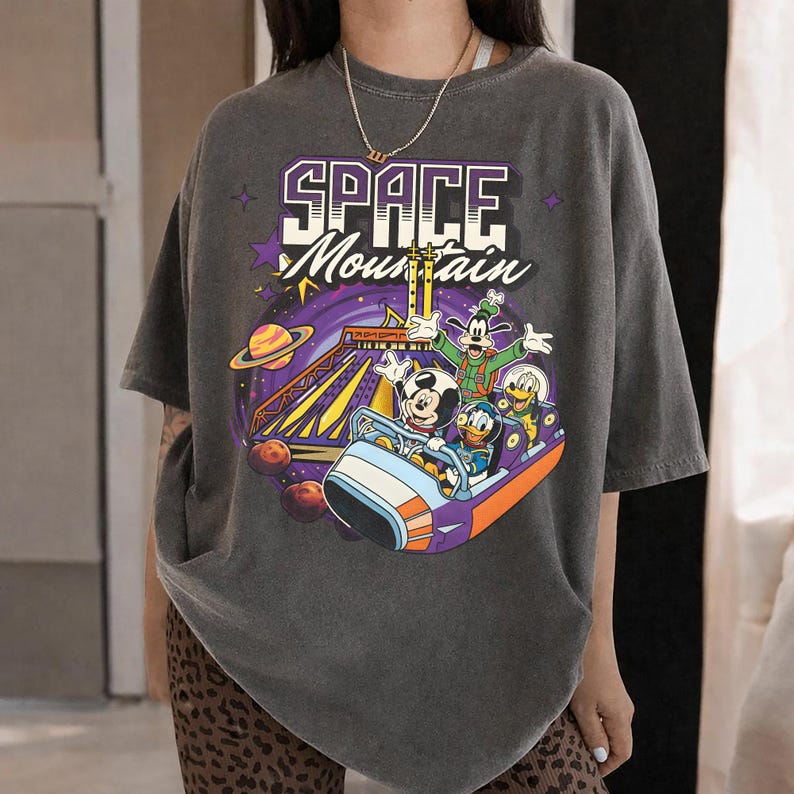 May include: Dark grey t-shirt with a colourful graphic of the Space Mountain ride featuring Mickey Mouse, Donald Duck, Goofy, and Pluto. The shirt has the words "SPACE Mountain" in a purple and white design.