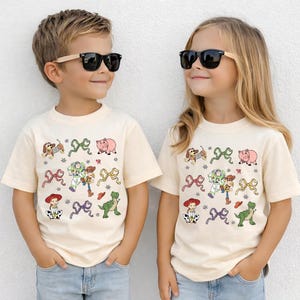 Op de afbeelding: Twee kinderen dragen bijpassende cr&egrave;mekleurige T-shirts met een kleurrijk cartoonontwerp. Het ontwerp bevat karakters en strikken. De shirts worden gecombineerd met een blauwe jeans en een zonnebril.