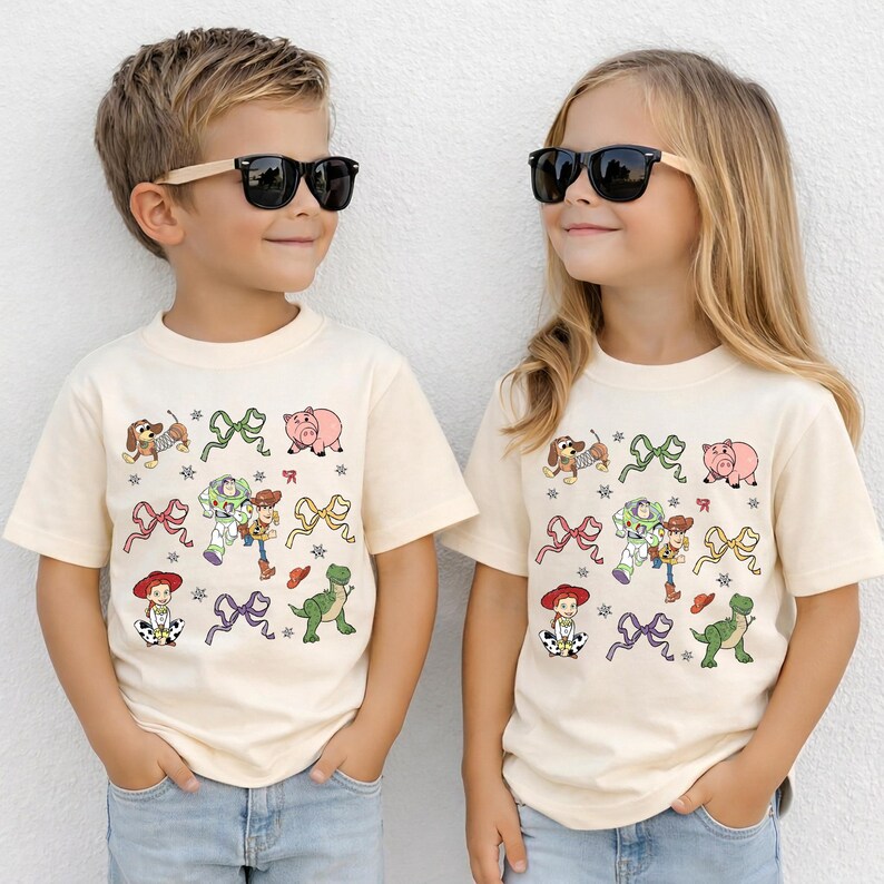 Op de afbeelding: Twee kinderen dragen bijpassende cr&egrave;mekleurige T-shirts met een kleurrijk cartoonontwerp. Het ontwerp bevat karakters en strikken. De shirts worden gecombineerd met een blauwe jeans en een zonnebril.