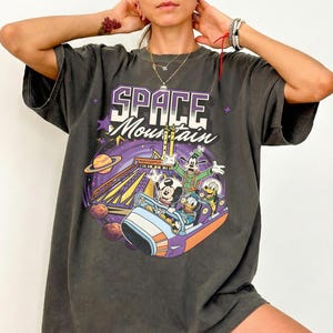 May include: Dark grey t-shirt with a Space Mountain graphic featuring Mickey Mouse, Goofy, and Donald Duck. The words "SPACE Mountain" are in purple and white. The t-shirt has an oversized fit.