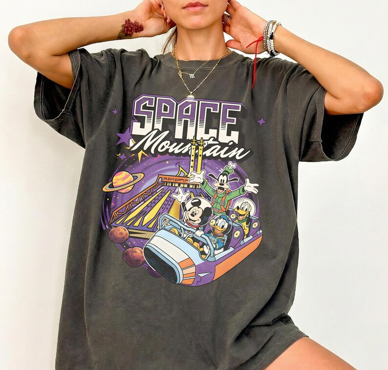 May include: Dark grey t-shirt with a Space Mountain graphic featuring Mickey Mouse, Goofy, and Donald Duck. The words "SPACE Mountain" are in purple and white. The t-shirt has an oversized fit.