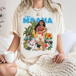 May include: Cream-colored t-shirt featuring a colorful graphic of a Polynesian woman, pig, and rooster, with palm trees and flowers. The text "I Am Moana" is printed in blue above the graphic. The shirt has short sleeves.