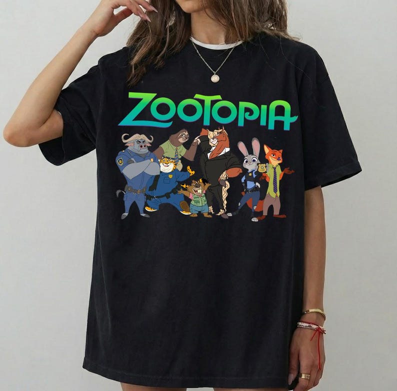 K&ouml;nnte beinhalten: Schwarzes T-Shirt mit dem Wort "ZOOTOPIA" in gr&uuml;nen und blauen Farbverlauf-Buchstaben. Das Shirt zeigt Zeichentrickfiguren aus dem Film, darunter einen Polizisten, ein Faultier und einen Fuchs.