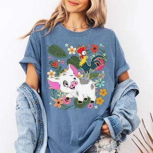 Puede incluir: Camiseta azul con un dise&ntilde;o de cerdo y gallo de dibujos animados, rodeada de flores y hojas coloridas. La camiseta tiene un corte holgado y mangas cortas, adecuada para el uso diario. El dise&ntilde;o es vibrante y colorido.