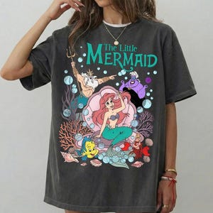 Puede incluir: Camiseta gris oscuro con un gr&aacute;fico colorido de personajes de "La Sirenita". El dise&ntilde;o incluye a Ariel, Flounder, Sebasti&aacute;n, el Rey Trit&oacute;n y &Uacute;rsula, con el t&iacute;tulo en verde azulado. La camiseta tiene un corte holgado.