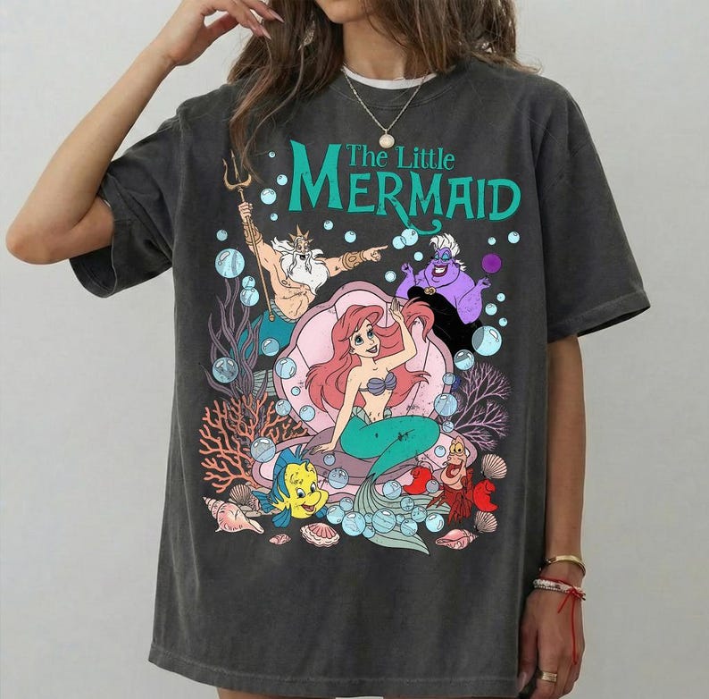 Puede incluir: Camiseta gris oscuro con un gr&aacute;fico colorido de personajes de "La Sirenita". El dise&ntilde;o incluye a Ariel, Flounder, Sebasti&aacute;n, el Rey Trit&oacute;n y &Uacute;rsula, con el t&iacute;tulo en verde azulado. La camiseta tiene un corte holgado.