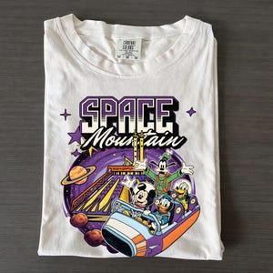 May include: White t-shirt with a graphic of Mickey Mouse, Donald Duck, and Goofy on a Space Mountain roller coaster. The design includes the words "SPACE MOUNTAIN" in purple and white, with stars and planets.