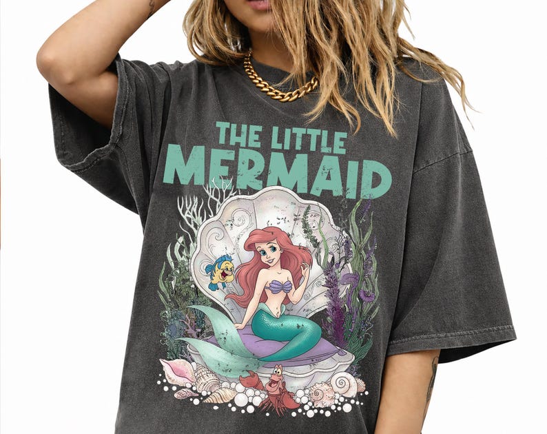 May include: Dark gray t-shirt featuring a graphic of Ariel from The Little Mermaid. The design includes Ariel in a seashell, with Flounder and Sebastian. The text "THE LITTLE MERMAID" is above the image in teal.