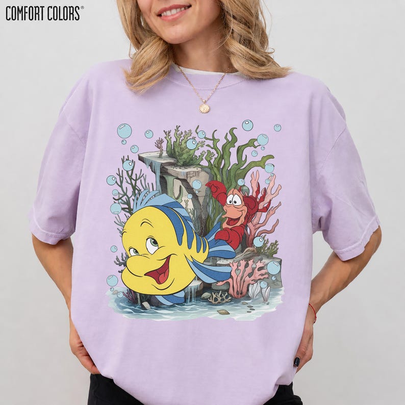 Puede incluir: Una camiseta de color lila claro con un dibujo animado de Flounder y Sebasti&aacute;n de La Sirenita. El dise&ntilde;o incluye plantas acu&aacute;ticas y burbujas. El texto "COMFORT COLORS" est&aacute; impreso en la parte superior izquierda.